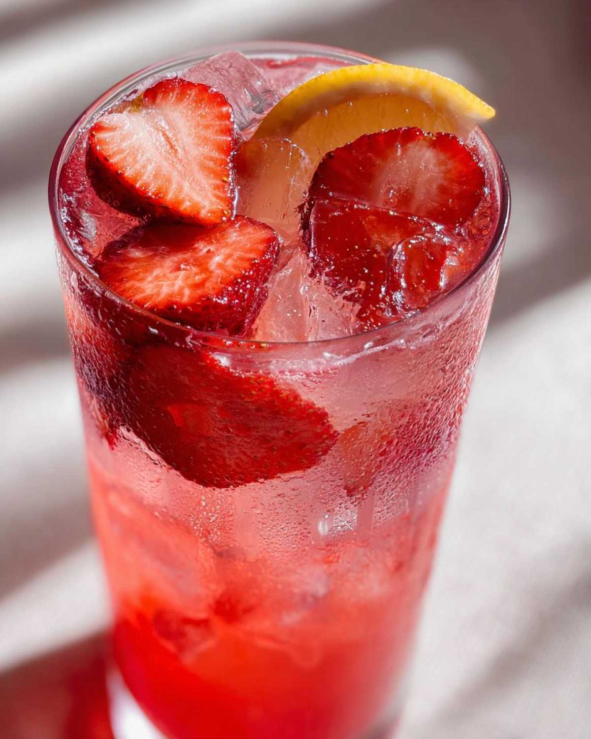 Close-up of a tall glass of vibrant red Iced Strawberry Lemon Tea filled with ice, strawberry slices, and a lemon wedge.