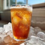 A tall glass filled with a bubbly, amber-colored Iced Vanilla Chai Latte, surrounded by large ice cubes.