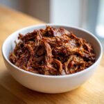 A white bowl filled with rich, shredded Instant Pot Beef Barbacoa, glistening with sauce.