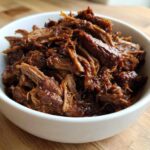 A white bowl filled with richly seasoned, shredded Instant Pot Beef Barbacoa resting on a wooden surface.