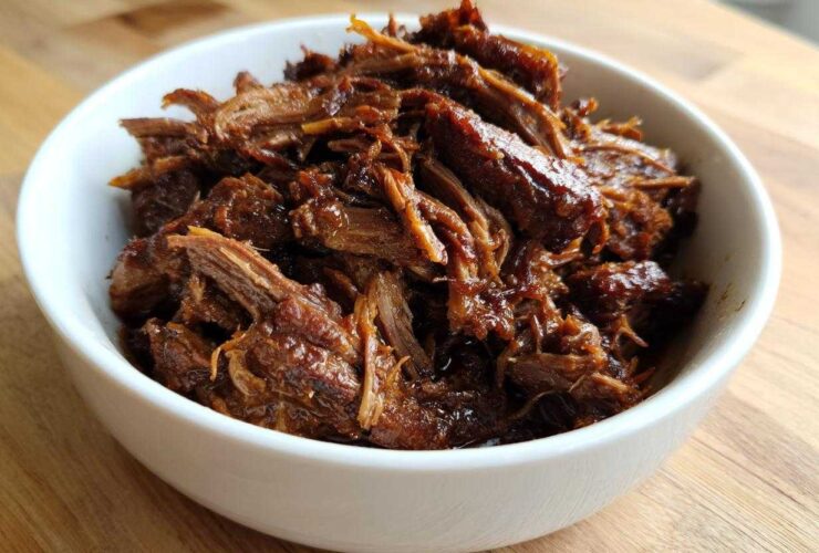 A white bowl filled with richly seasoned, shredded Instant Pot Beef Barbacoa resting on a wooden surface.