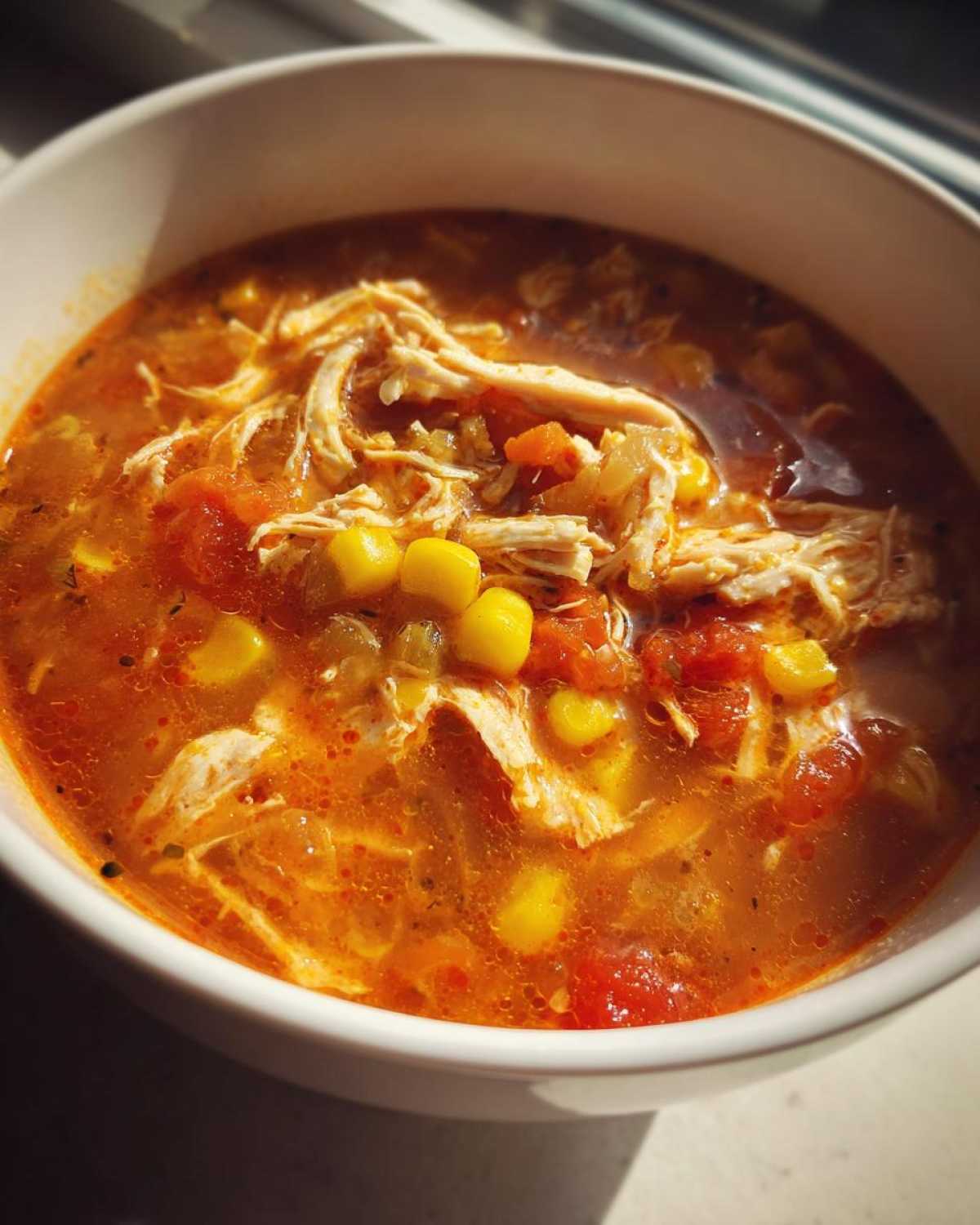 Close-up of a white bowl filled with rich, orange broth Instant Pot Chicken Tortilla Soup, featuring shredded chicken and bright yellow corn kernels.