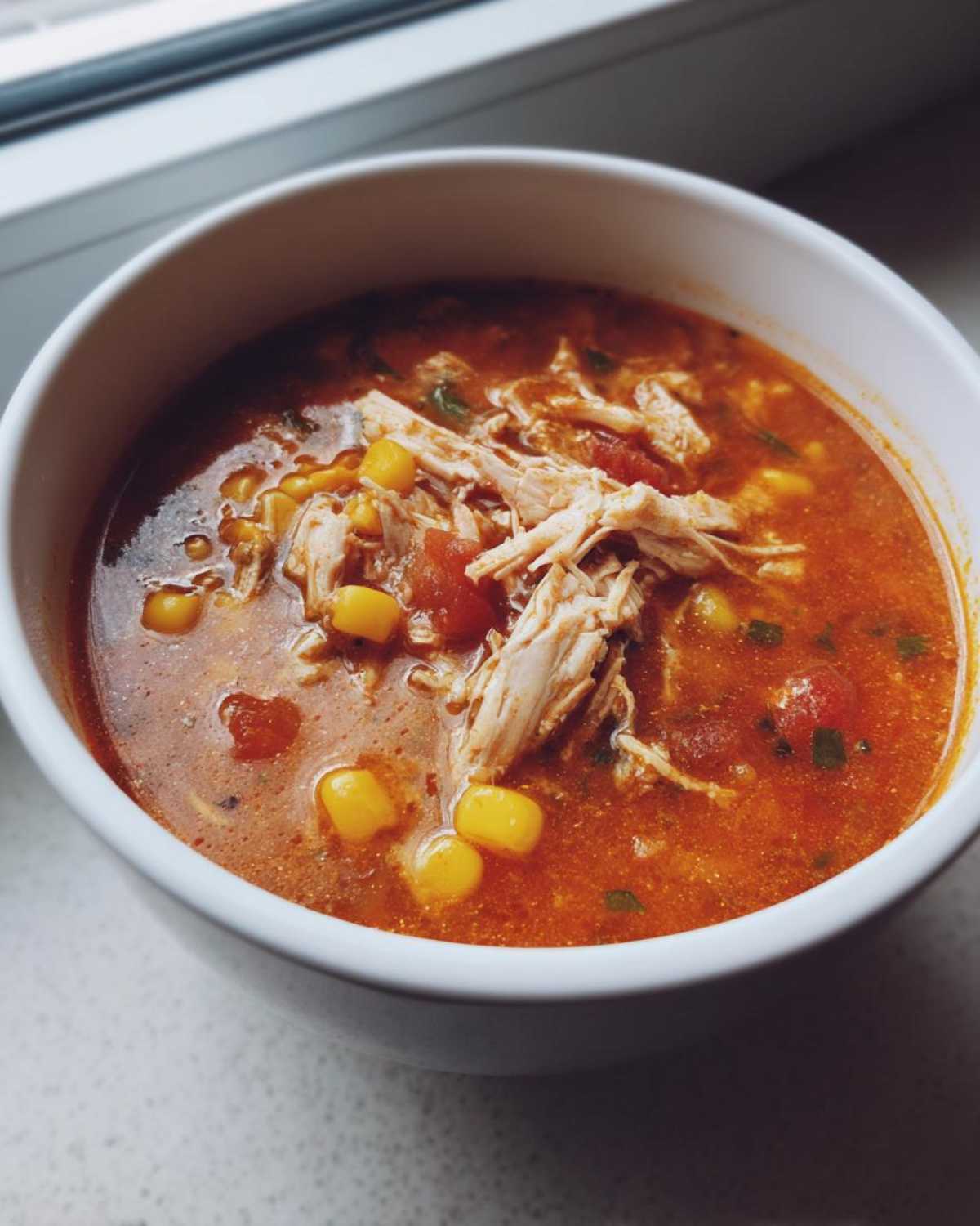 A close-up of a white bowl filled with rich, red Instant Pot Chicken Tortilla Soup, topped with shredded chicken and corn kernels.