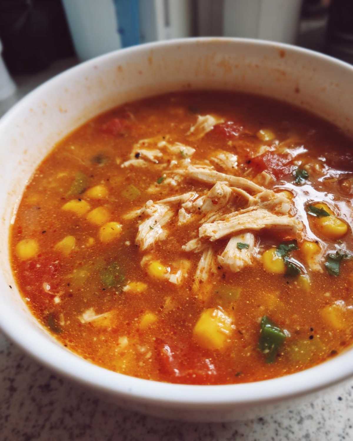 A close-up view of a white bowl filled with rich, orange-red Instant Pot Chicken Tortilla Soup, featuring shredded chicken, corn, and tomatoes.