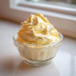 A serving of light yellow, fluffy Keto Lemon Cheesecake Mousse piped high in a small clear glass bowl.