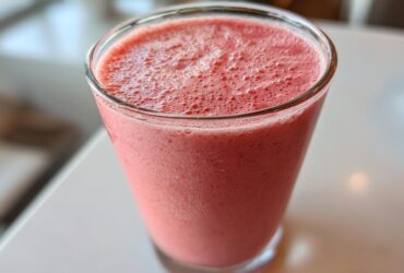 Close-up of a tall glass filled with a thick, pink Keto Strawberry Cheesecake Smoothie, showing a frothy top.