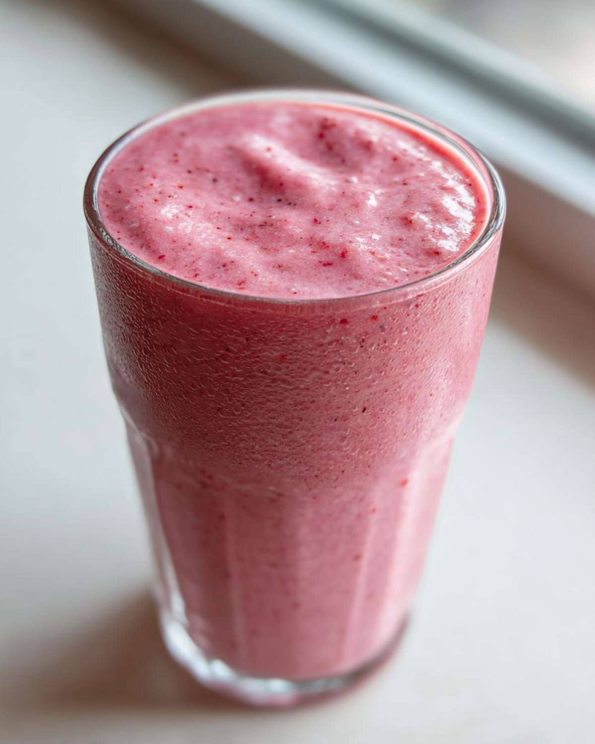 Close-up of a tall glass filled with a thick, pink Keto Strawberry Cheesecake Smoothie showing condensation on the glass.