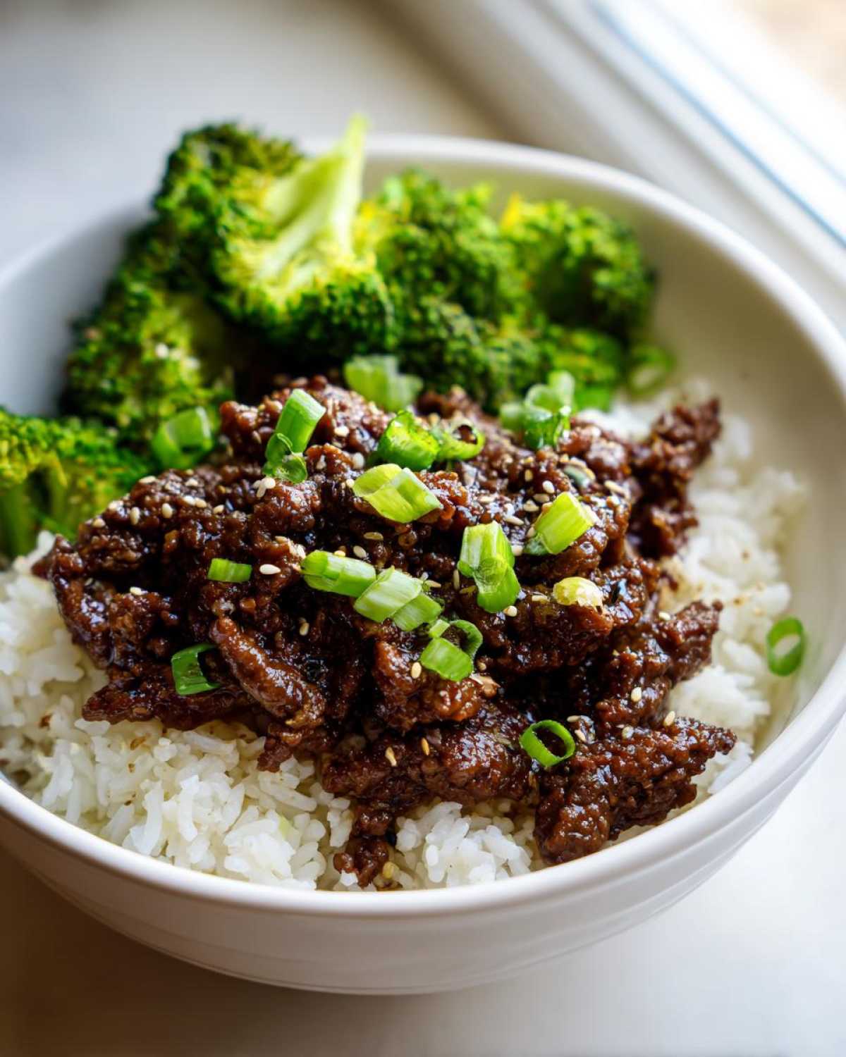 Close-up of Korean Ground Beef Bulgogi Bowls featuring saucy ground beef over white rice, topped with green onions and sesame seeds, served with steamed broccoli.