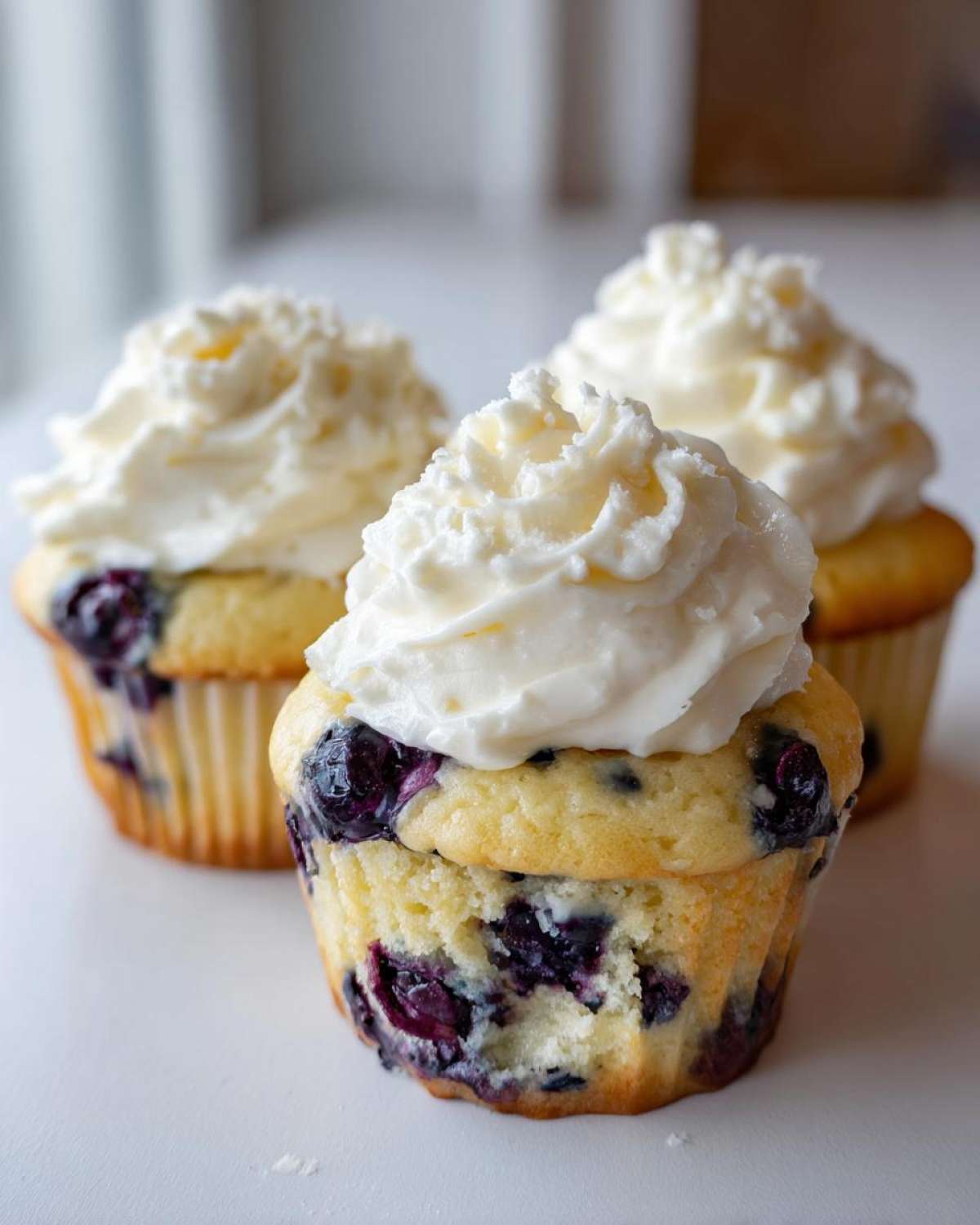 Three Lemon Blueberry Cupcakes topped with swirls of white cream cheese frosting on a white surface.