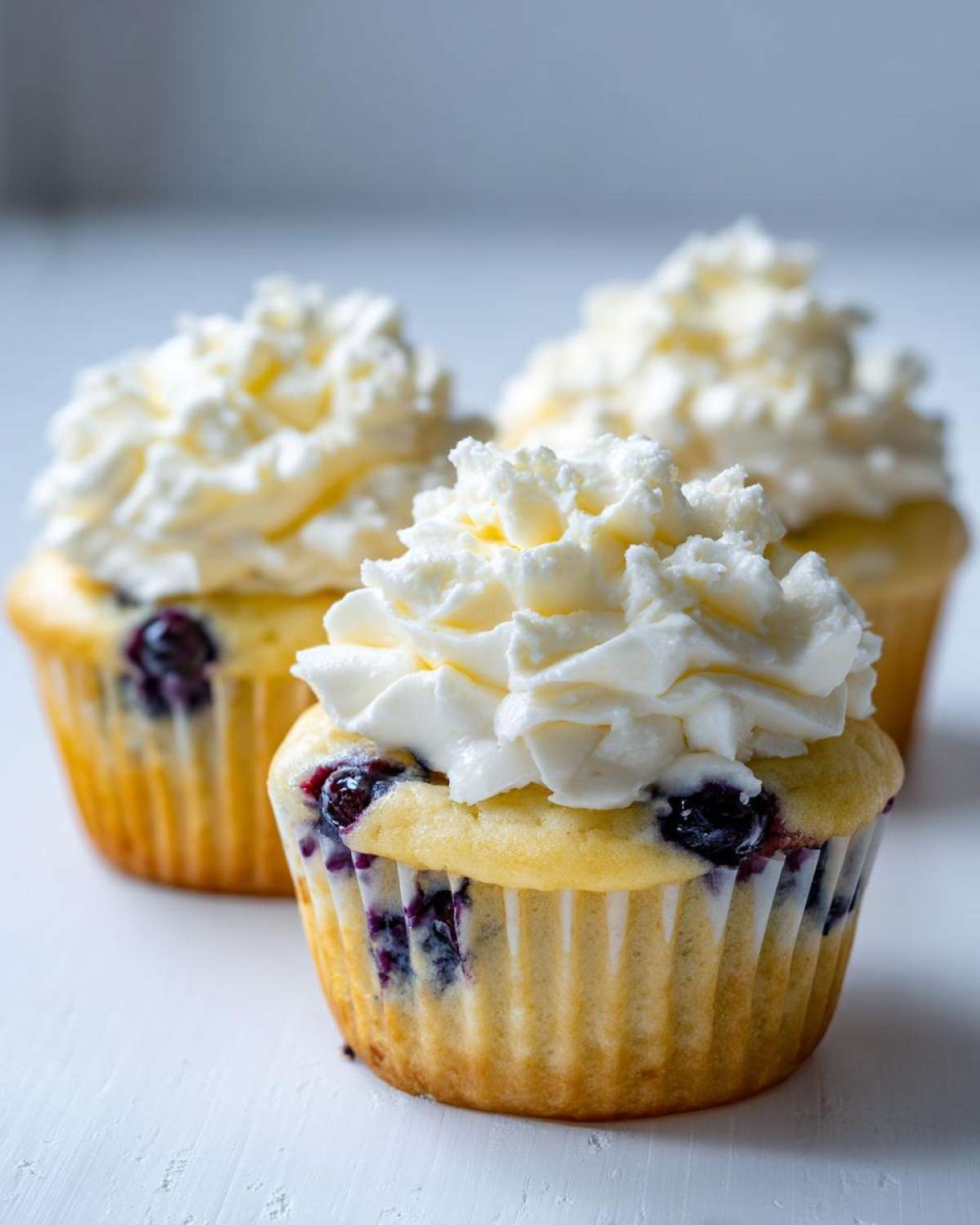 Three Lemon Blueberry Cupcakes topped with generous swirls of cream cheese frosting on a white surface.