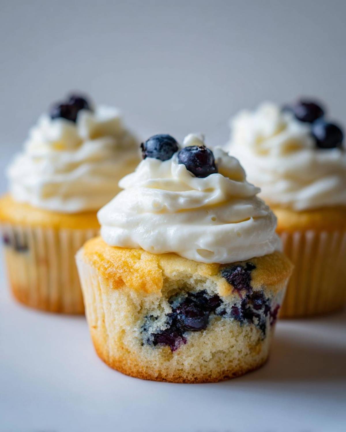 A close-up of a Lemon Blueberry Cupcake with a bite taken out, showing moist cake and blueberries, topped with cream cheese frosting.