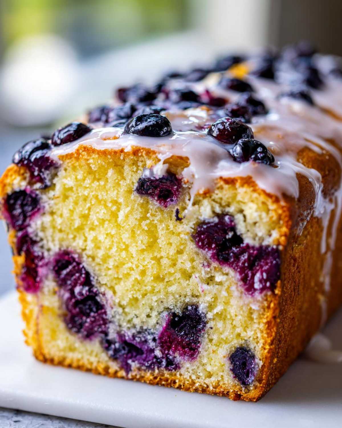 Close-up of a slice of moist Lemon Blueberry Yogurt Loaf topped with fresh blueberries and a white glaze.