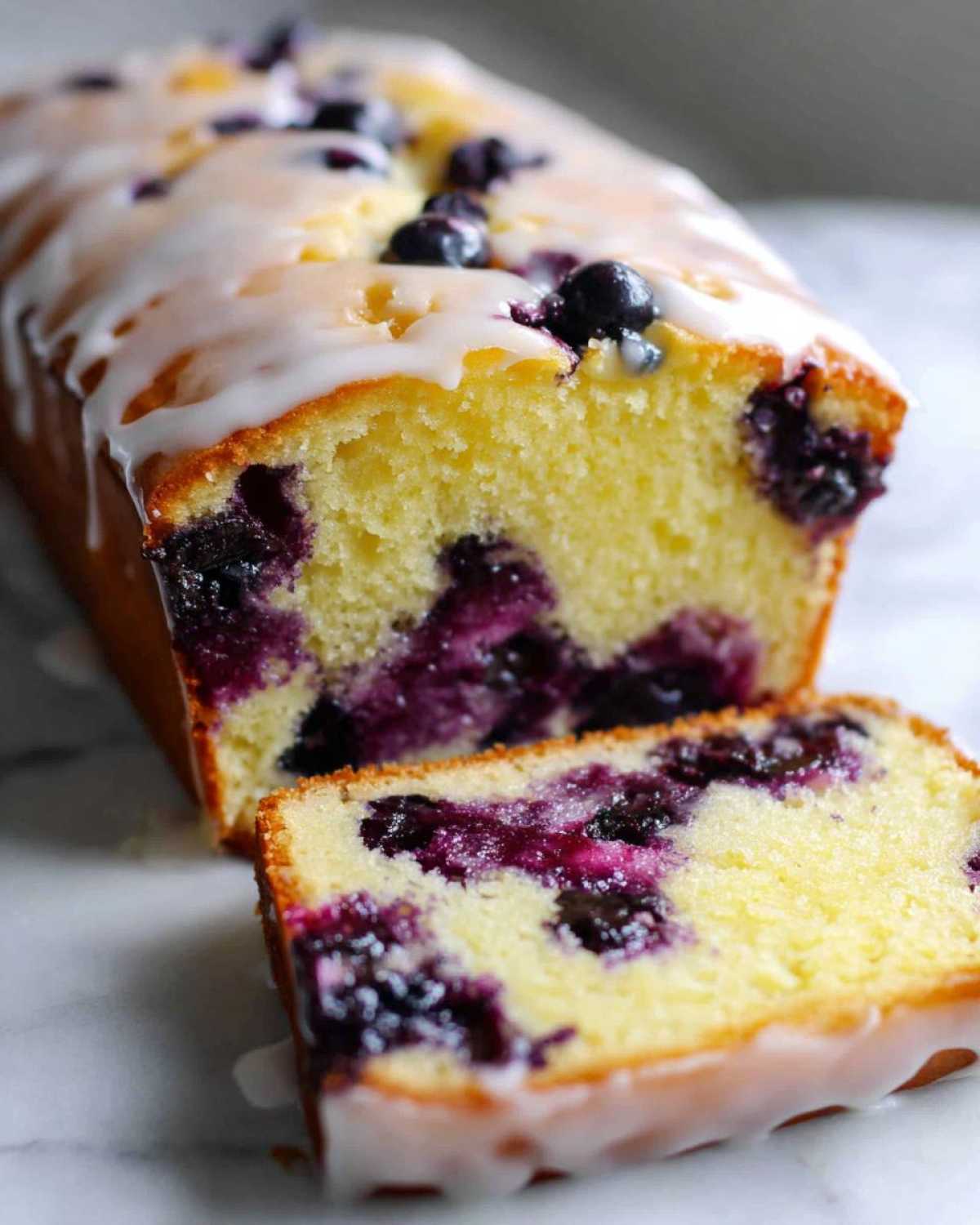 Close-up of a moist Lemon Blueberry Yogurt Loaf, sliced to show blueberries and topped with a white glaze.