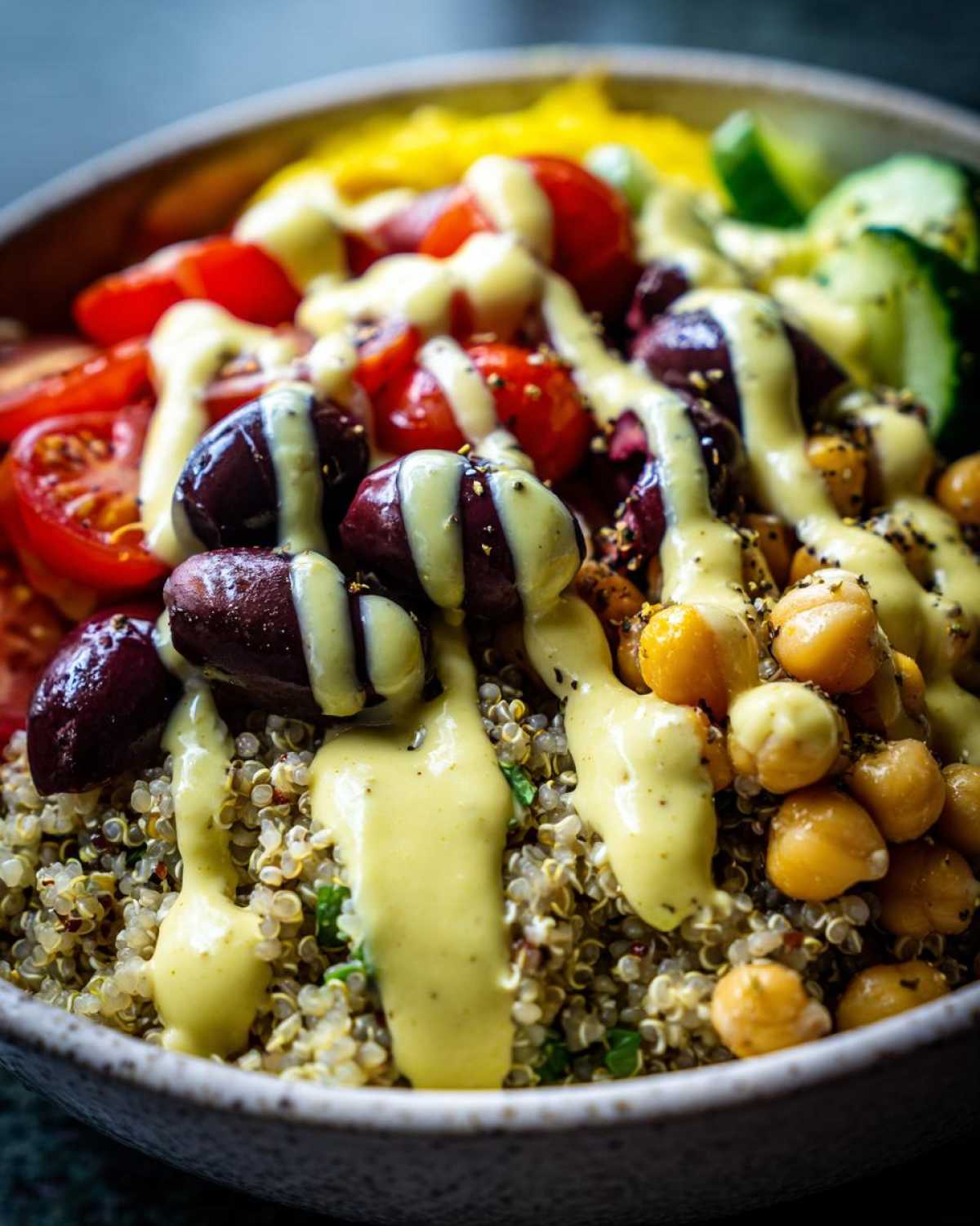Close-up of a Mediterranean Quinoa Bowl topped with olives, chickpeas, tomatoes, and a generous drizzle of lemon tahini sauce.