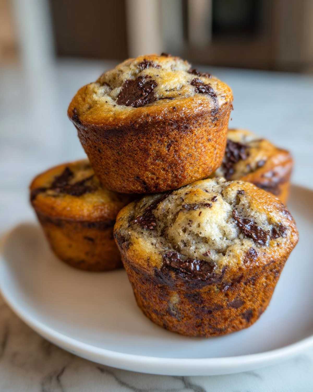 A stack of four moist Chocolate Chip Banana Muffins with melted chocolate chunks on a white plate.