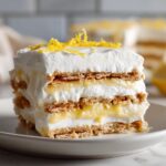 A tall slice of No-Bake Lemon Icebox Cake featuring layers of graham crackers, lemon filling, and whipped topping, garnished with zest.