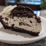 A generous slice of No-Bake Oreo Cheesecake on a white plate, topped with whipped cream and Oreo crumbs.