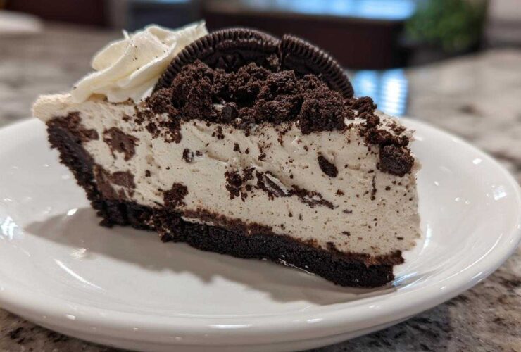 A generous slice of No-Bake Oreo Cheesecake on a white plate, topped with whipped cream and Oreo crumbs.