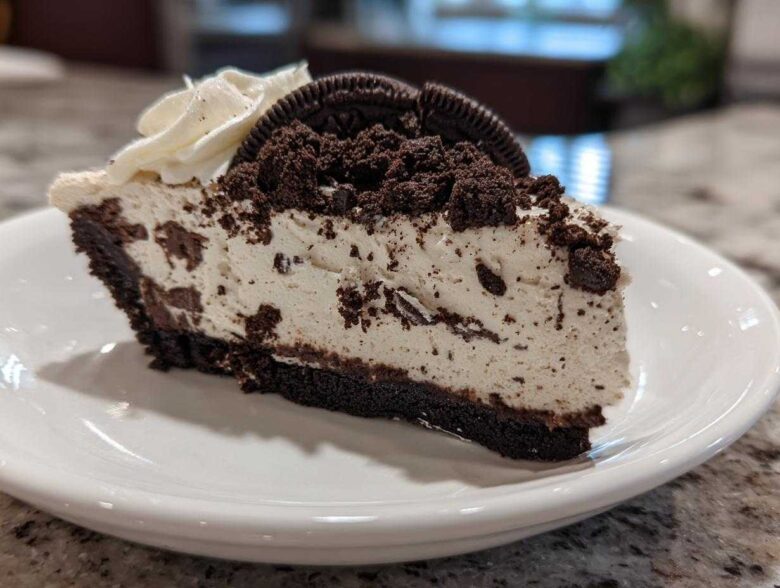 A generous slice of No-Bake Oreo Cheesecake on a white plate, topped with whipped cream and Oreo crumbs.
