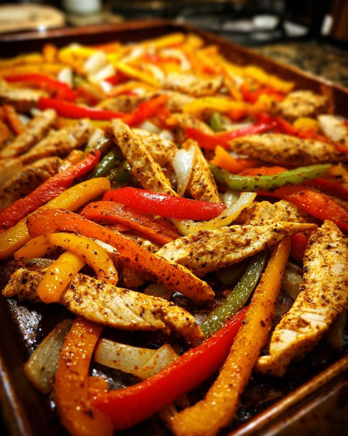 Close-up of seasoned chicken strips, red, yellow, and green peppers, and onions cooked together on a sheet pan for One-Pan Chicken Fajitas.