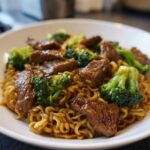 A close-up shot of a white bowl filled with glossy noodles, tender beef pieces, and bright green broccoli florets from the One-Pot Beef and Broccoli Ramen Noodles.