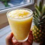 A hand holding a glass filled with a bright yellow, icy Pineapple Coconut Smoothie, with a whole pineapple in the background.