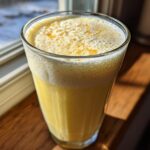 A tall glass filled with a bright yellow Pineapple Coconut Smoothie, topped with foam, sitting on a wooden windowsill.