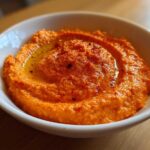Close-up of vibrant orange Roasted Red Pepper Hummus swirled in a white bowl, drizzled with olive oil and paprika.