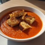 A bowl of vibrant Roasted Tomato Basil Soup topped with golden Grilled Cheese Croutons, seasoned with pepper.