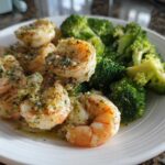 A white plate featuring cooked shrimp generously coated in garlic butter sauce next to bright green broccoli florets.