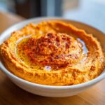 Close-up of vibrant orange Roasted Red Pepper Hummus, swirled and topped with olive oil and paprika.