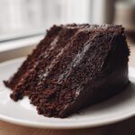 A close-up of a moist, dark slice of Gluten-Free Chocolate Cake covered in rich frosting on a white plate.