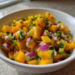Close-up of a white bowl filled with bright orange chunks of mango, red onion, and cilantro, representing Spicy Mango Salsa.