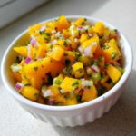 Close-up of bright orange Spicy Mango Salsa with diced red onion and cilantro in a white bowl.