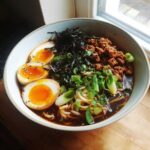 A close-up bowl of Spicy Miso Ramen featuring rich broth, noodles, ground meat, green onions, nori, and perfectly halved soft-boiled eggs.