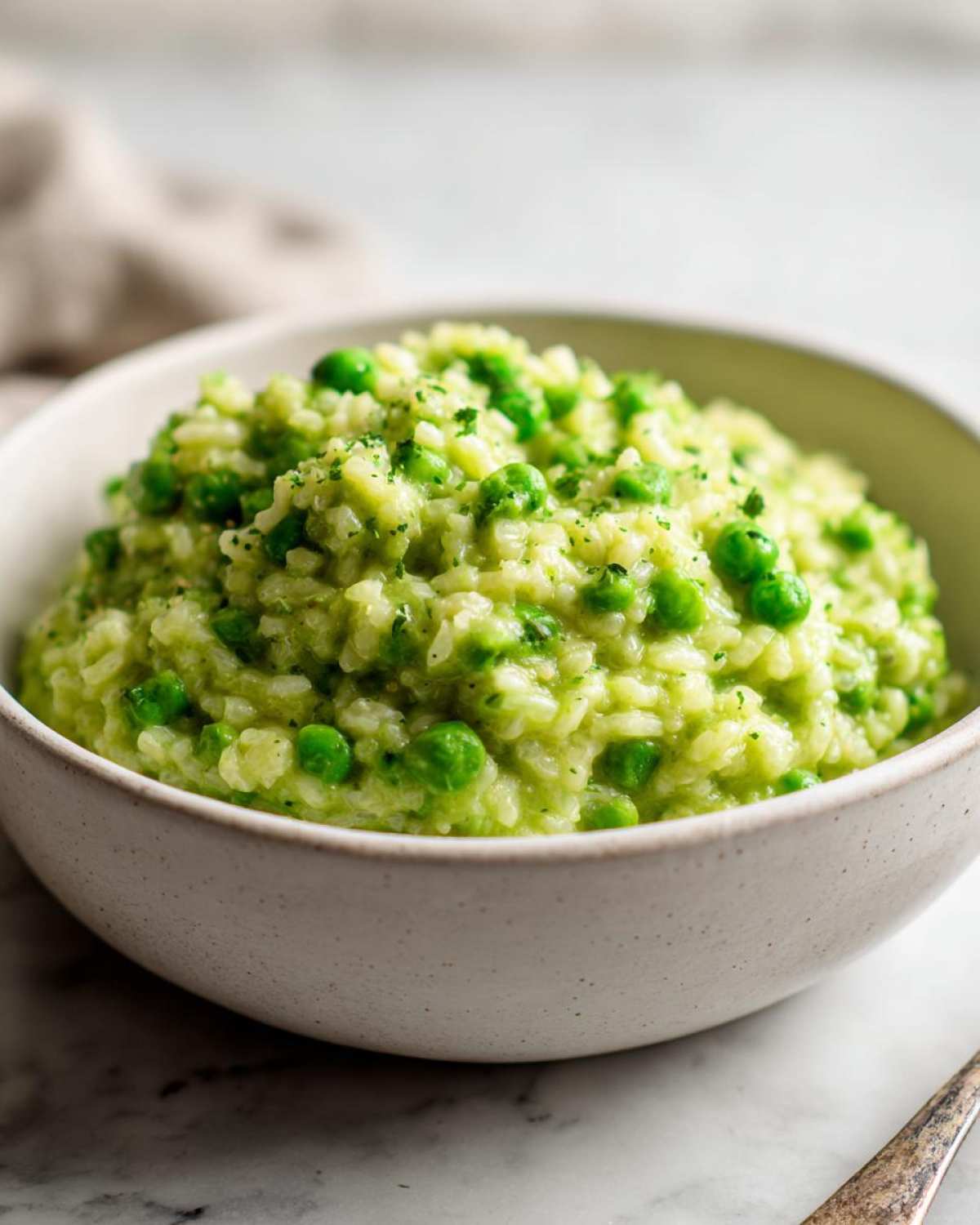 A close-up of creamy Spring Pea and Mint Risotto topped with whole peas and fresh herbs in a light ceramic bowl.