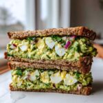 A tall, stacked Avocado Egg Salad Sandwich featuring creamy green filling, visible hard-boiled egg slices, red onion, and herbs on whole wheat bread.
