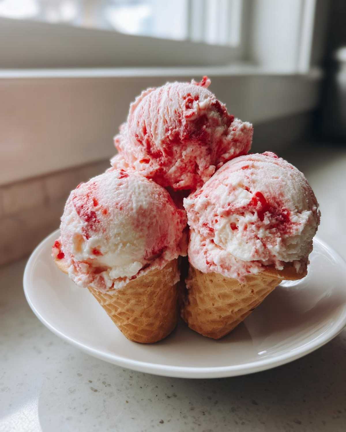 Three scoops of swirled pink and white Strawberry Banana Ice Cream served in small waffle cones on a white plate.