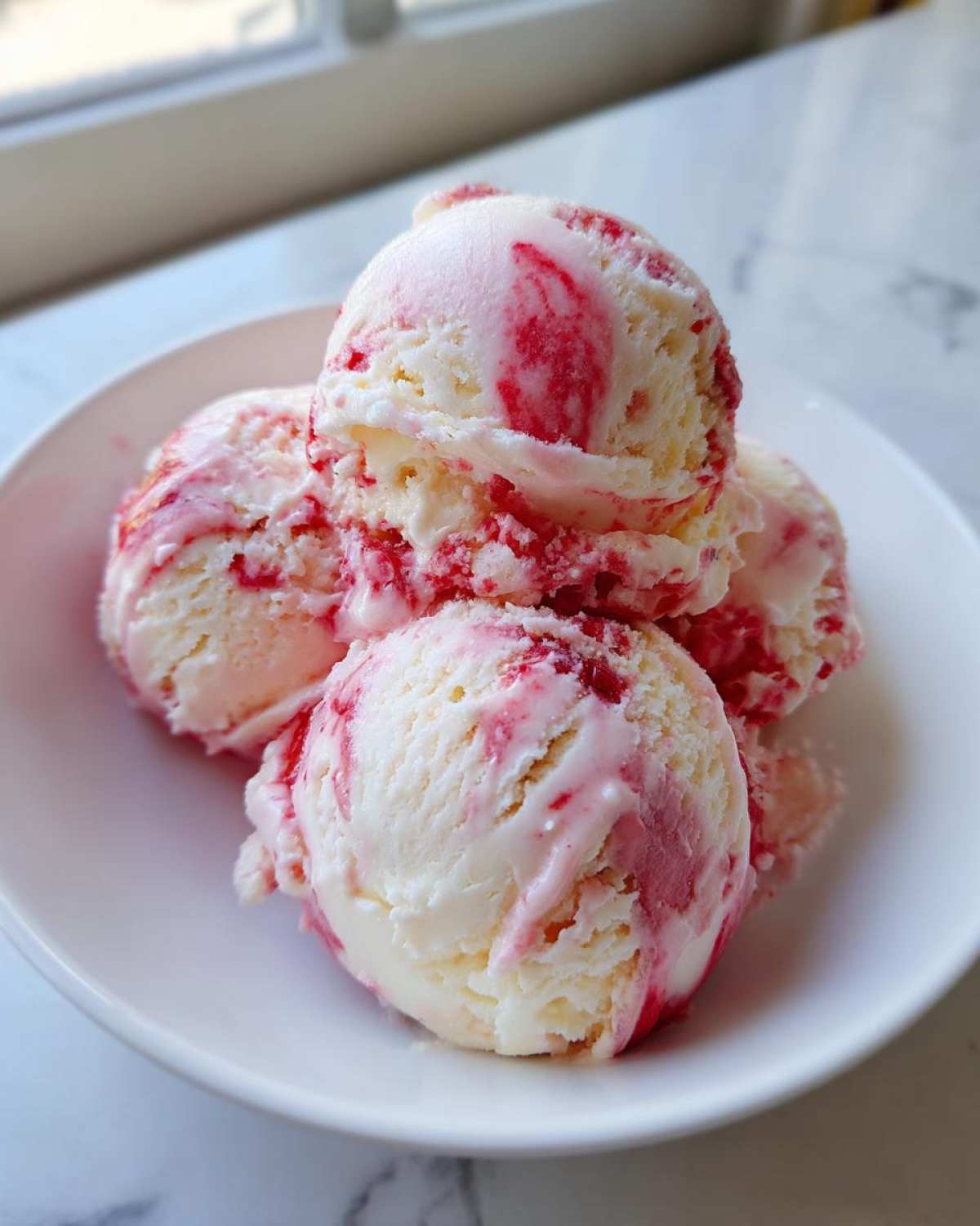 Four scoops of creamy Strawberry Banana Ice Cream with visible red strawberry swirls served in a white bowl.