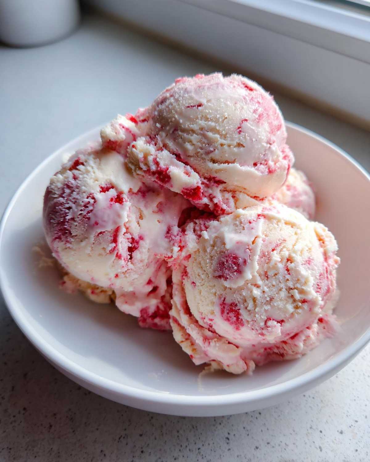 Close-up of several scoops of homemade Strawberry Banana Ice Cream swirled with pink fruit pieces in a white bowl.