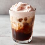 A tall glass filled with ice, dark cold brew coffee, and topped with thick, pink strawberry cream foam.