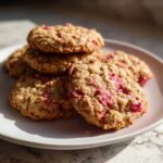 Close-up of several textured Strawberry Oatmeal Breakfast Cookies stacked on a pale plate, featuring visible oats and bright pink strawberry pieces.