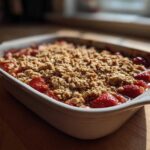 A close-up of a freshly baked Strawberry Rhubarb Crisp with a golden oat topping in a white baking dish.