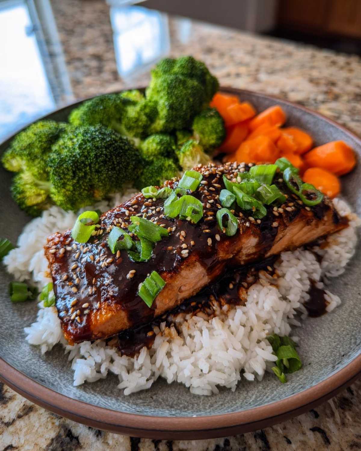 Close-up of a Teriyaki Salmon Rice Bowl featuring glazed salmon over white rice, broccoli, and carrots, topped with sesame seeds and scallions.