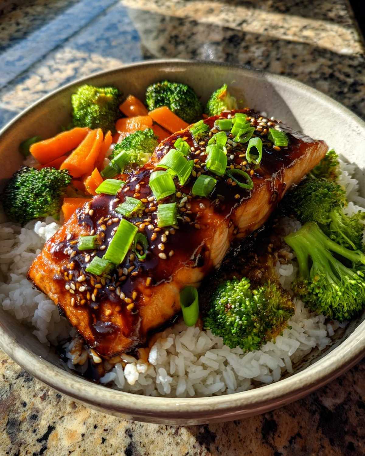 A close-up of a Teriyaki Salmon Rice Bowl featuring a glazed salmon fillet over white rice with steamed broccoli and carrots.