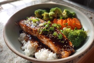 A close-up of a Teriyaki Salmon Rice Bowls featuring glazed salmon over white rice with steamed broccoli and sliced carrots.