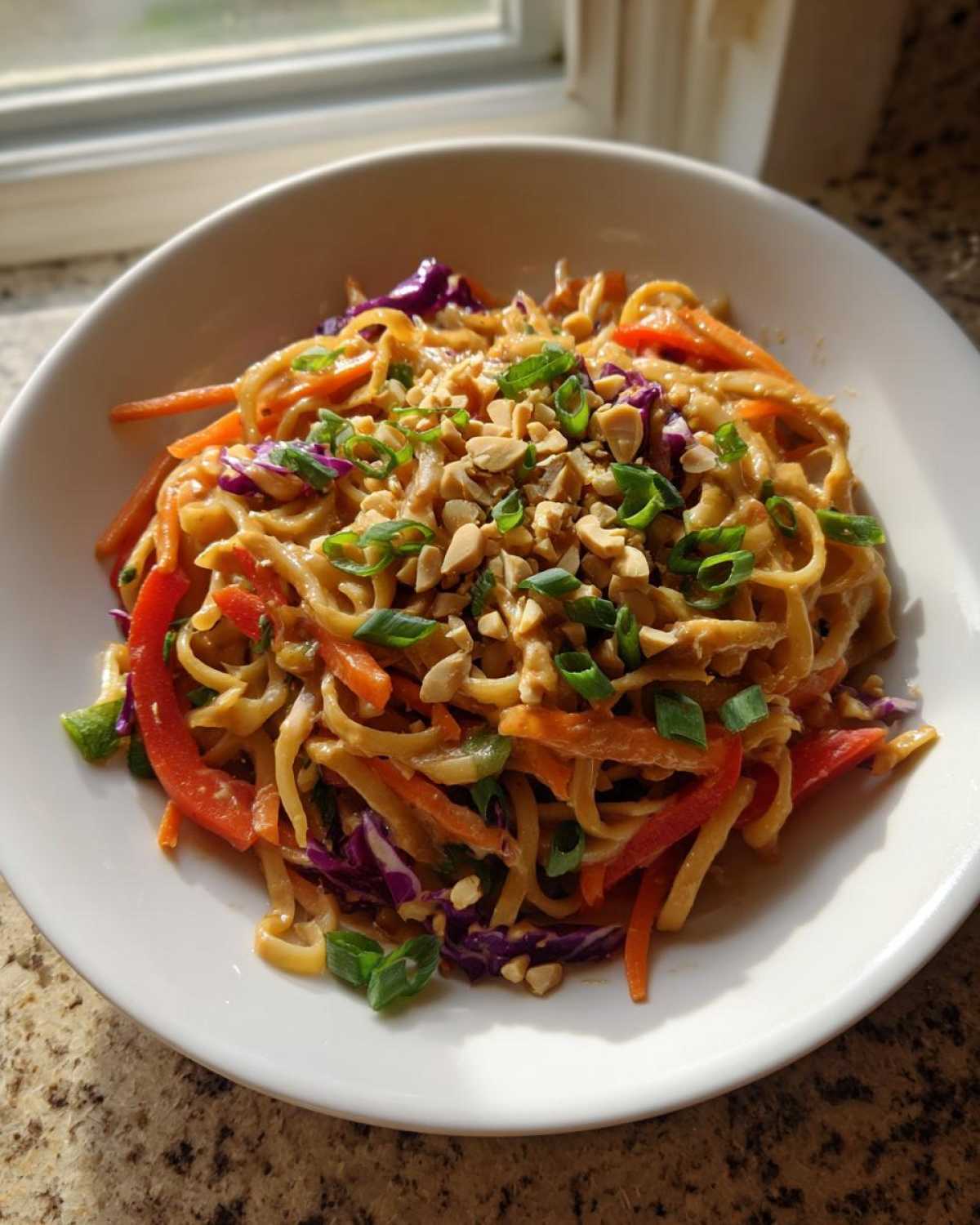 A white bowl filled with saucy Thai Peanut Noodles with Crunchy Veggies, topped with chopped peanuts and green onions.