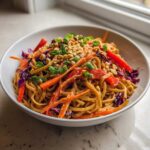 A close-up of a white bowl filled with creamy Thai Peanut Noodles with Crunchy Veggies, topped with peanuts and scallions.