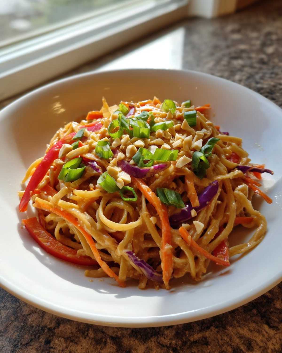 A close-up of Thai Peanut Noodles with Crunchy Veggies, topped with peanuts and green onions in a white bowl.