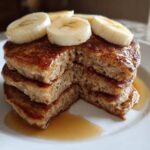 A close-up stack of fluffy Vegan Banana Oat Pancakes drizzled with syrup and topped with fresh banana slices.